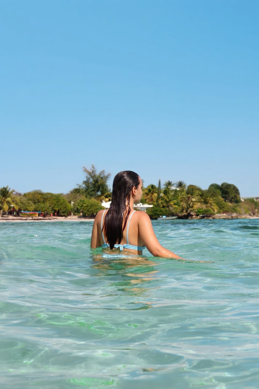 Marta de costas, dentro de água transparente, a olhar para uma praia com palmeiras em Guadalupe