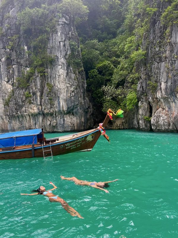 Duas pessoas a flutuar em água verde-esmeralda junto a um barco long-tail na Tailândia